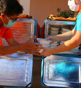 Foto de um homem entregando para outro três marmitas, e ao fundo há duas mochilas térmicas e uma delas está aberta e seu interior está cheio de marmitas
