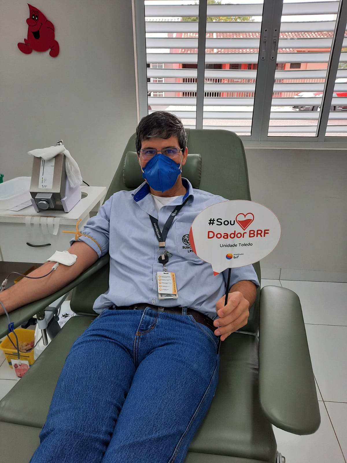 Foto de um homem deitado em uma cadeira enquanto está doando sangue. Ele segura uma placa com o texto #SouDoadorBRF Unidade Toledo.
