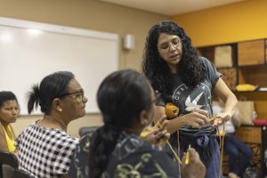 Na foto, uma mulher vestindo uma blusa cinza com um desenho branco está de pé. Ela mostra algo para outras duas mulheres, que estão sentadas