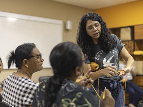 Na foto, uma mulher vestindo uma blusa cinza com um desenho branco está de pé. Ela mostra algo para outras duas mulheres, que estão sentadas
