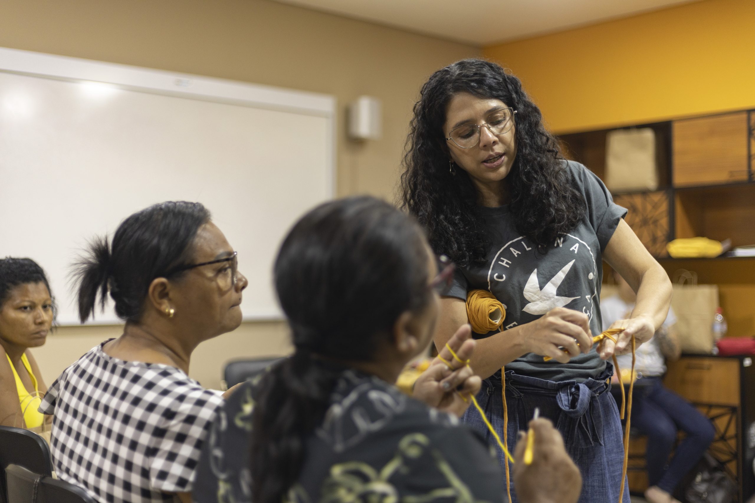 Na foto, uma mulher vestindo uma blusa cinza com um desenho branco está de pé. Ela mostra algo para outras duas mulheres, que estão sentadas